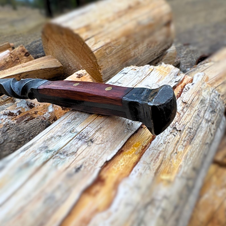 May include: A hand-forged knife with a dark wooden handle and a twisted metal blade. The handle is secured with brass rivets. The knife rests on a pile of wooden logs, suggesting a rustic or outdoor theme.