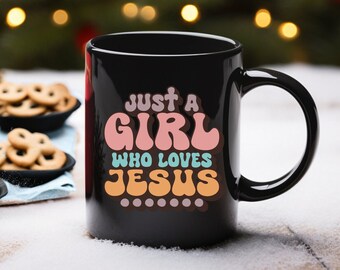 Op de afbeelding: Zwarte keramische koffiemok met een wit ontwerp met de tekst "Just a Girl Who Loves Jesus" in kleurrijke, retro-stijl letters.