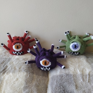 May include: Three crocheted Beholder monsters, one red, one purple, and one green, with a single large eye and multiple tentacles.
