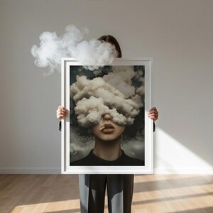 May include: A person holding a framed art print. The print features a woman's face obscured by a cloud of smoke, with a cigarette in her mouth. The artwork is in a white frame, and the background is a neutral wall and wooden floor.