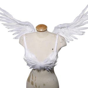 May include: A white feather bra and matching feathered wings are displayed on a mannequin. The bra has a delicate design, and the wings are spread wide. The mannequin has a wooden head and a cream-colored torso.
