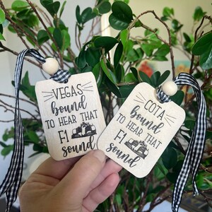 May include: Two wooden tags with black and white checkered ribbon. The tags are decorated with a black and white illustration of a race car and the text "Vegas bound to hear that F1 sound" and "COTA bound to hear that F1 sound".