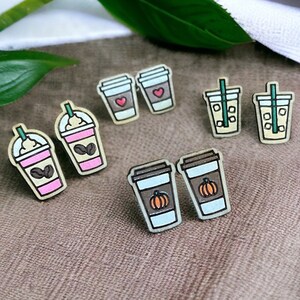 May include: Six pairs of wooden earrings shaped like coffee cups. The earrings feature different designs, including a pumpkin spice latte, a regular coffee, a latte with a heart, and a iced coffee with a straw.