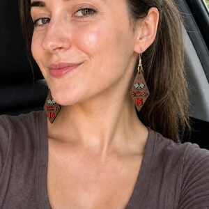 May include: Wooden diamond-shaped earrings with a racing theme. The earrings are brown with red and white accents, featuring a checkered flag design and the number 16. The earrings are hanging from gold-colored hooks.