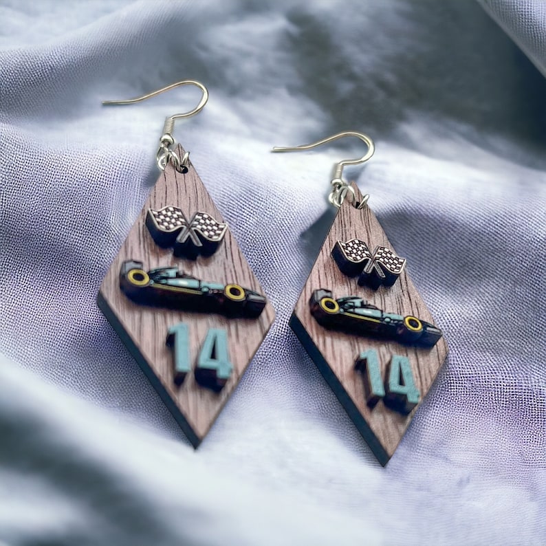 May include: A pair of diamond-shaped earrings with a wood grain pattern. Each earring features a race car, checkered flags, and the number 14 in teal. The earrings have silver-colored fish hook closures.