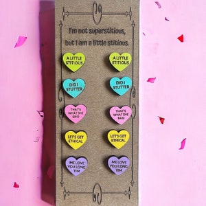 May include: A brown card with the text "I'm not superstitious, but I am a little stitious."  The card has eight heart-shaped candy-like decorations with various sayings on them, including "A Little Stitious", "Did I Stutter", "That's What She Said", "Let's Get Ethical", and "Me Love You Long Tim".