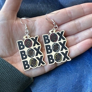 May include: A pair of wooden earrings with the word "BOX" repeated four times on each earring. The word "BOX" is in black lettering with a red, yellow, and black circle design inside each letter. The earrings are hanging from silver hooks.