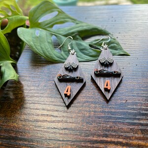 May include: A pair of wooden earrings with a racing theme. Each earring features a dark brown, diamond-shaped base with a silver hook. The earrings have a checkered flag, a race car, and the number 4 in orange.