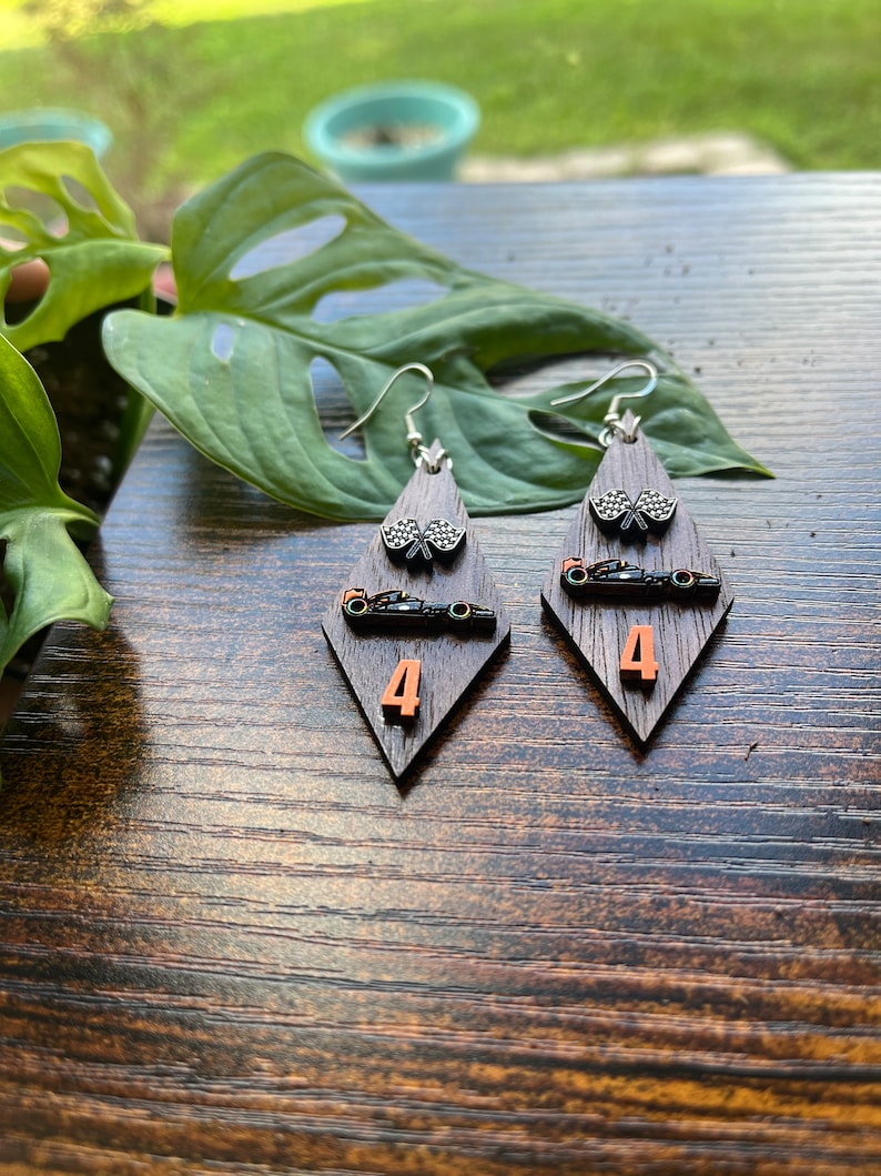May include: A pair of wooden earrings with a racing theme. Each earring features a dark brown, diamond-shaped base with a silver hook. The earrings have a checkered flag, a race car, and the number 4 in orange.