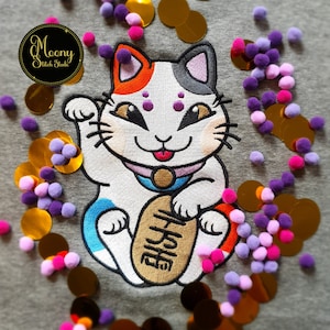 May include: An embroidered lucky cat with white, grey and orange fur, holding a gold coin in its paw. The cat is holding a gold sign with Japanese characters. The cat is surrounded by gold sequins and purple and pink pom-poms.