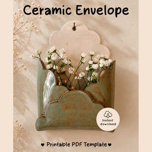 Peut inclure: Une pochette murale en céramique vert sauge en forme d'enveloppe à bord festonné, contenant des fleurs blanches de gypsophile. Le texte "Ceramic Envelope" figure en haut et "Printable PDF Template" en bas avec une icône de téléchargement.