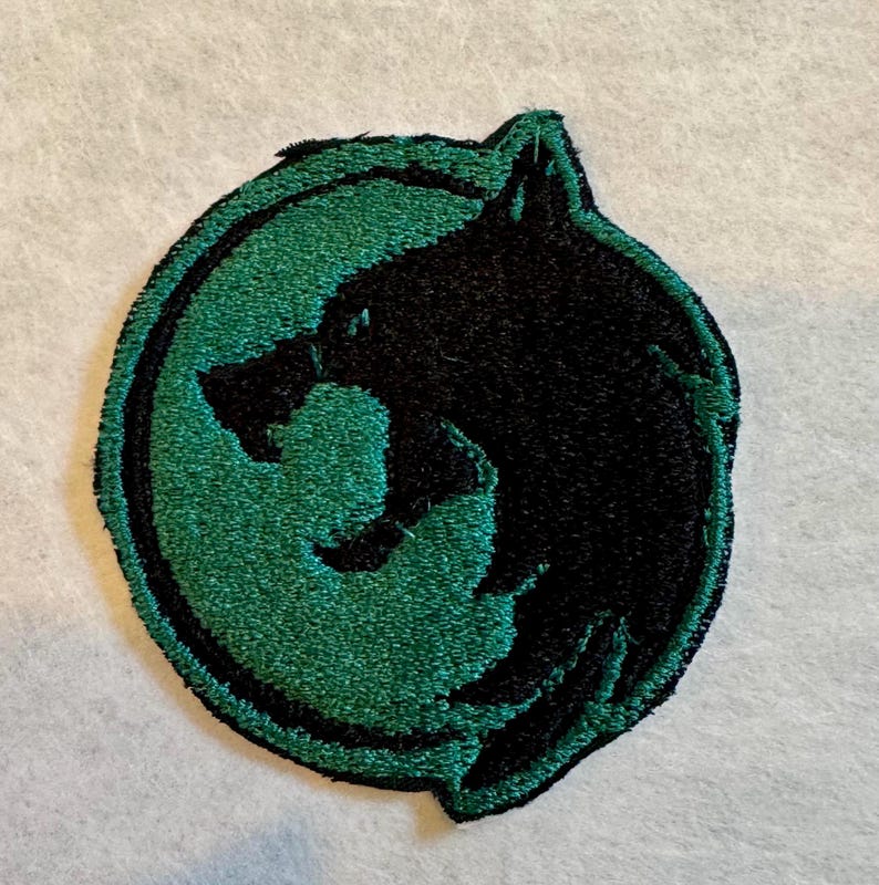 K&ouml;nnte beinhalten: Gestickter Aufn&auml;her mit einer schwarzen Wolfskopf-Silhouette in einem gr&uuml;nen Kreis. Der Aufn&auml;her hat eine strukturierte Oberfl&auml;che, wobei sich der Wolfskopf vom gr&uuml;nen Hintergrund abhebt. Der Aufn&auml;her ist wahrscheinlich f&uuml;r Kleidung oder Accessoires gedacht.