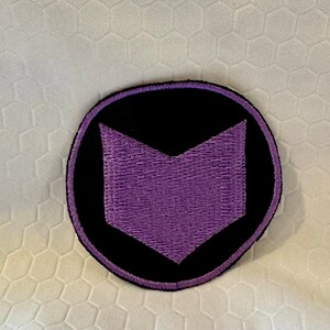 May include: A purple and black embroidered patch with a purple arrow shape in the center. The patch is circular and has a black background.