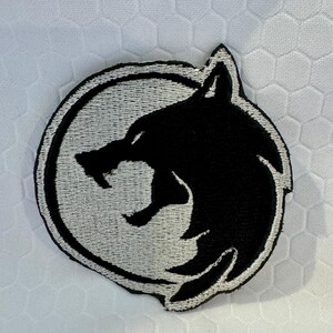 K&ouml;nnte beinhalten: Schwarz-wei&szlig;er, gestickter Aufn&auml;her mit einem Wolfskopf in einem Kreis. Der Wolf ist im Profil mit offenem Maul dargestellt. Der Aufn&auml;her befindet sich auf einem wei&szlig;en, strukturierten Hintergrund, geeignet f&uuml;r Kleidung oder Accessoires.