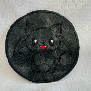 Puede incluir: Un parche bordado en blanco y negro de un murciélago de dibujos animados con la lengua roja. El murciélago está sobre un fondo circular negro.