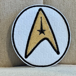 May include: A round, white patch with a black border and a gold embroidered Starfleet insignia. The insignia is a stylised, pointed shape with a five-pointed star in the centre.