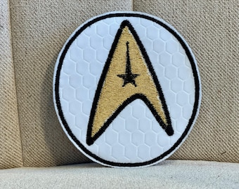 Könnte beinhalten: Ein runder, weißer Patch mit schwarzem Rand und einem goldenen gestickten Starfleet-Abzeichen. Das Abzeichen ist eine stilisierte, spitze Form mit einem fünfzackigen Stern in der Mitte.