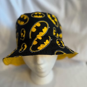 May include: A black bucket hat with a yellow Batman logo pattern. The hat is on a white mannequin head.