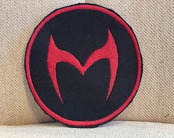 Könnte beinhalten: Ein schwarzer runder Patch mit rotem Rand und einem roten gestickten Buchstaben M.
