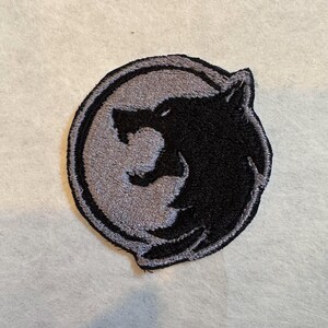 K&ouml;nnte beinhalten: Gestickter Aufn&auml;her mit einem schwarzen Wolfskopf-Design auf grauem Hintergrund, umgeben von einem kreisf&ouml;rmigen Rand. Der Aufn&auml;her ist f&uuml;r Kleidung oder Accessoires geeignet und bietet ein dekoratives Element mit Naturthema.