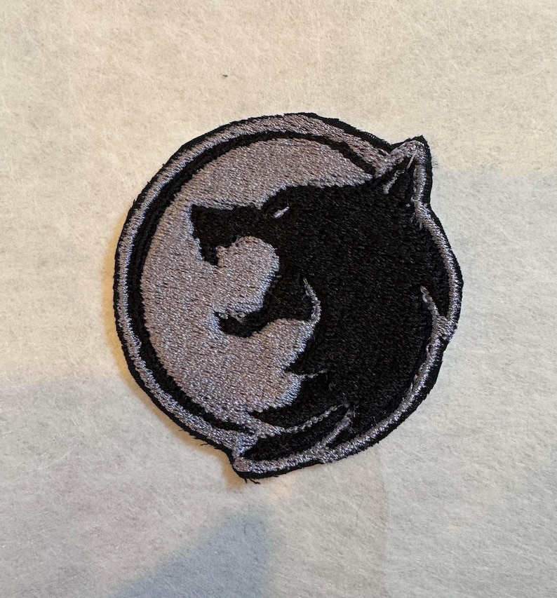 K&ouml;nnte beinhalten: Gestickter Aufn&auml;her mit einem schwarzen Wolfskopf-Design auf grauem Hintergrund, umgeben von einem kreisf&ouml;rmigen Rand. Der Aufn&auml;her ist f&uuml;r Kleidung oder Accessoires geeignet und bietet ein dekoratives Element mit Naturthema.