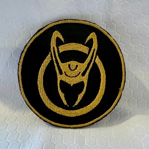 Könnte beinhalten: Ein gestickter Aufnäher in Schwarz und Gold mit einem stilisierten Helm mit Hörnern. Der Helm ist in Gold umrandet und hat einen goldenen Kreis darum.