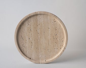 May include: A round, beige stone tray with a raised edge. The tray has a natural, textured surface with small holes and variations in color. The neutral color and simple design make it suitable for various uses.