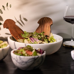 May include: A white marble salad bowl with a green salad and wooden salad servers. There are three smaller white marble bowls with red and white sprinkles, salt, and a gold spoon.