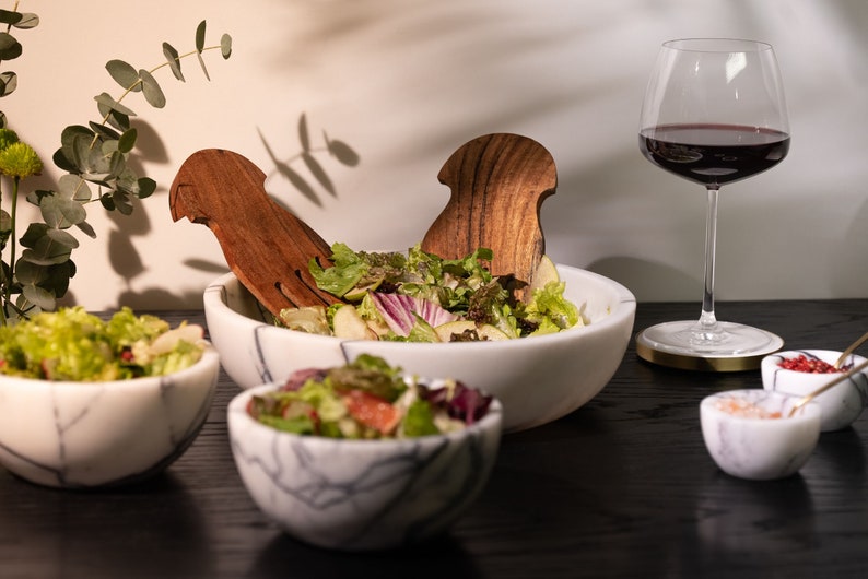May include: A white marble salad bowl with a green salad and wooden salad servers. There are three smaller white marble bowls with red and white sprinkles, salt, and a gold spoon.