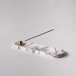 Könnte beinhalten: Weihrauchhalter aus weißem Marmor mit einer goldenen Metallkugel und einem schwarzen Weihrauchstäbchen. Der Halter ist oval geformt und hat eine glatte Oberfläche.