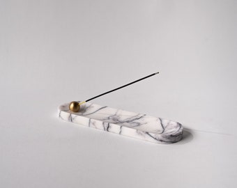 May include: White marble incense holder with a gold metal ball and a black incense stick. The holder is oval shaped and has a smooth surface.