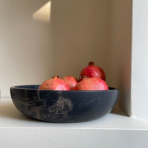 May include: A black marble bowl filled with red pomegranates. The bowl has a rustic, handmade look and is sitting on a white surface.