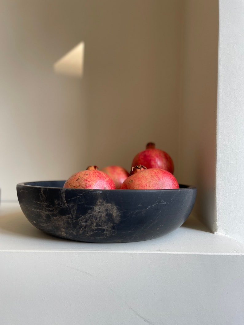 May include: A black marble bowl filled with red pomegranates. The bowl has a rustic, handmade look and is sitting on a white surface.