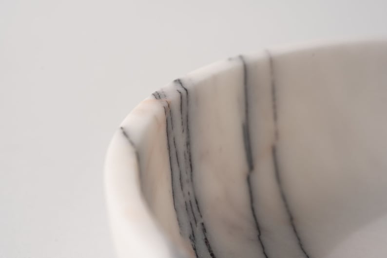 May include: Close-up of a white marble bowl with black veining. The bowl is smooth and polished, with a slightly curved edge.