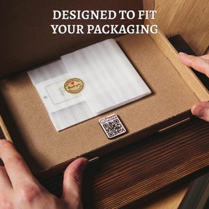 Op de afbeelding: Een bruine kartonnen doos met de tekst "DESIGNED TO FIT YOUR PACKAGING". Binnenin zit een witte schuimvellen, een kleine sticker met "thank you" en een sticker met een QR-code met de tekst "Please review".