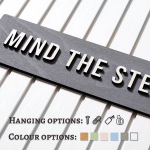 May include: A grey wooden sign with the text "MIND THE STEP" in white raised letters. The sign has hanging options including screws, rope, glue, and tape. The sign is available in six colours: brown, green, pink, blue, grey, and white.