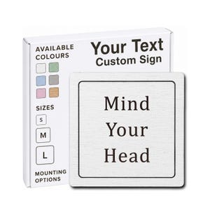 May include: A square white custom sign with a black border and the black text "Mind Your Head" centered on the surface. Behind the sign is a white product box labeled "Your Text Custom Sign" with icons for available colors, sizes S, M, L, and mounting options.