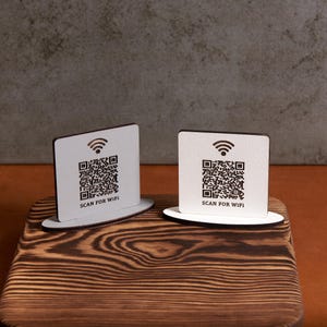 May include: Two white square signs with black QR codes and the text "SCAN FOR WIFI" on a wooden surface.