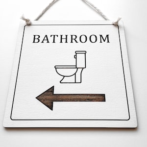 Op de afbeelding: Een wit houten bord met zwarte tekst die "Badkamer" luidt en een zwart silhouet van een toilet. Een zwarte pijl wijst naar links.