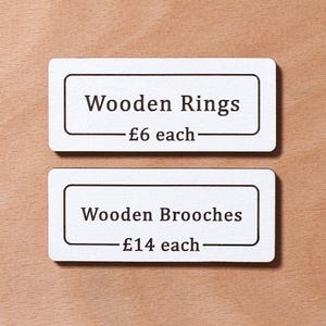 May include: Two white rectangular signs with black borders and text. The top sign reads "Wooden Rings - £6 each". The bottom sign reads "Wooden Brooches - £14 each".
