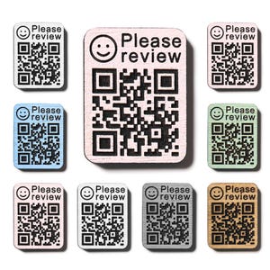 Op de afbeelding: Een verzameling houten QR-codetags in verschillende kleuren, waaronder wit, blauw, roze, groen, grijs en bruin. Elke tag heeft een QR-code, een smiley-icoon en de tekst "Please review".