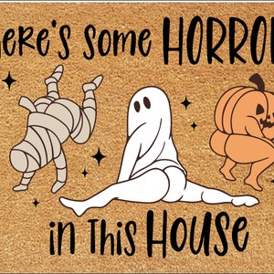Pu&ograve; includere: Zerbino rettangolare con sfondo beige e testo nero che recita "There's some HORRORS in This HOUSE." Il tappetino presenta illustrazioni di cartoni animati di una mummia, un fantasma e una zucca.