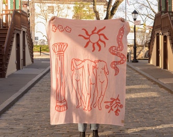 May include: A pink throw blanket with a graphic design featuring three figures, a snake, a sun, and a column. The figures are outlined in pink and have a peach-coloured interior.