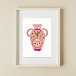 Può includere: Un vaso rosa con un disegno floreale dorato e un viso di sole al centro. Il vaso è incorniciato in una cornice bianca.