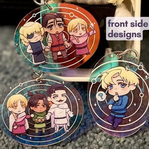 Peut inclure: Trois porte-clés ronds en acrylique avec des personnages de style anime. Chaque porte-clés présente un groupe différent de personnages aux couleurs vives, avec un fermoir en métal argenté. Le texte "front side designs" est visible.