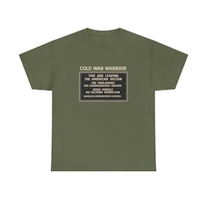 Könnte beinhalten: Olivgrünes T-Shirt mit dem Text "COLD WAR WARRIOR" über einem Schild mit der Aufschrift "YOU ARE LEAVING THE AMERICAN SECTOR" auf Englisch, Deutsch, Französisch und Russisch. Ein lässiges Kleidungsstück.
