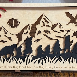 Puede incluir: Obra de arte de madera cortada con láser que representa una cordillera, un dragón y figuras. La pieza está hecha de madera de color claro con detalles en marrón oscuro y negro. El texto "One Ring to rule them all..." está en la parte inferior. Una figura roja con forma de dragón está en la parte superior izquierda.