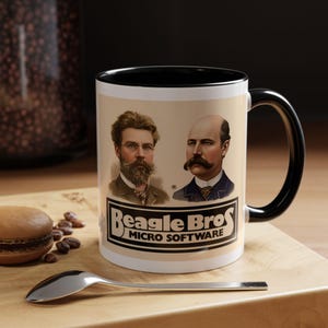 Puede incluir: Una taza de cerámica blanca con interior y asa negros. La taza presenta retratos de dos hombres y el texto "Beagle Bros MICRO SOFTWARE" en un diseño en blanco y negro. La taza está sobre una superficie de madera.