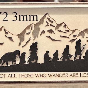 May include: A wooden frame with a laser-cut design of a mountain range and a group of figures from The Lord of the Rings. The text "V2 3mm" is visible in the top left corner. The text "Not all those who wander are lost" is visible at the bottom of the frame.