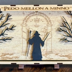 May include: A wooden shadow box with a silhouette of a figure holding a staff standing in front of a large archway. The archway is decorated with a star pattern and the text "PEDO MELLON A MINNO". The shadow box is on a wooden stand.